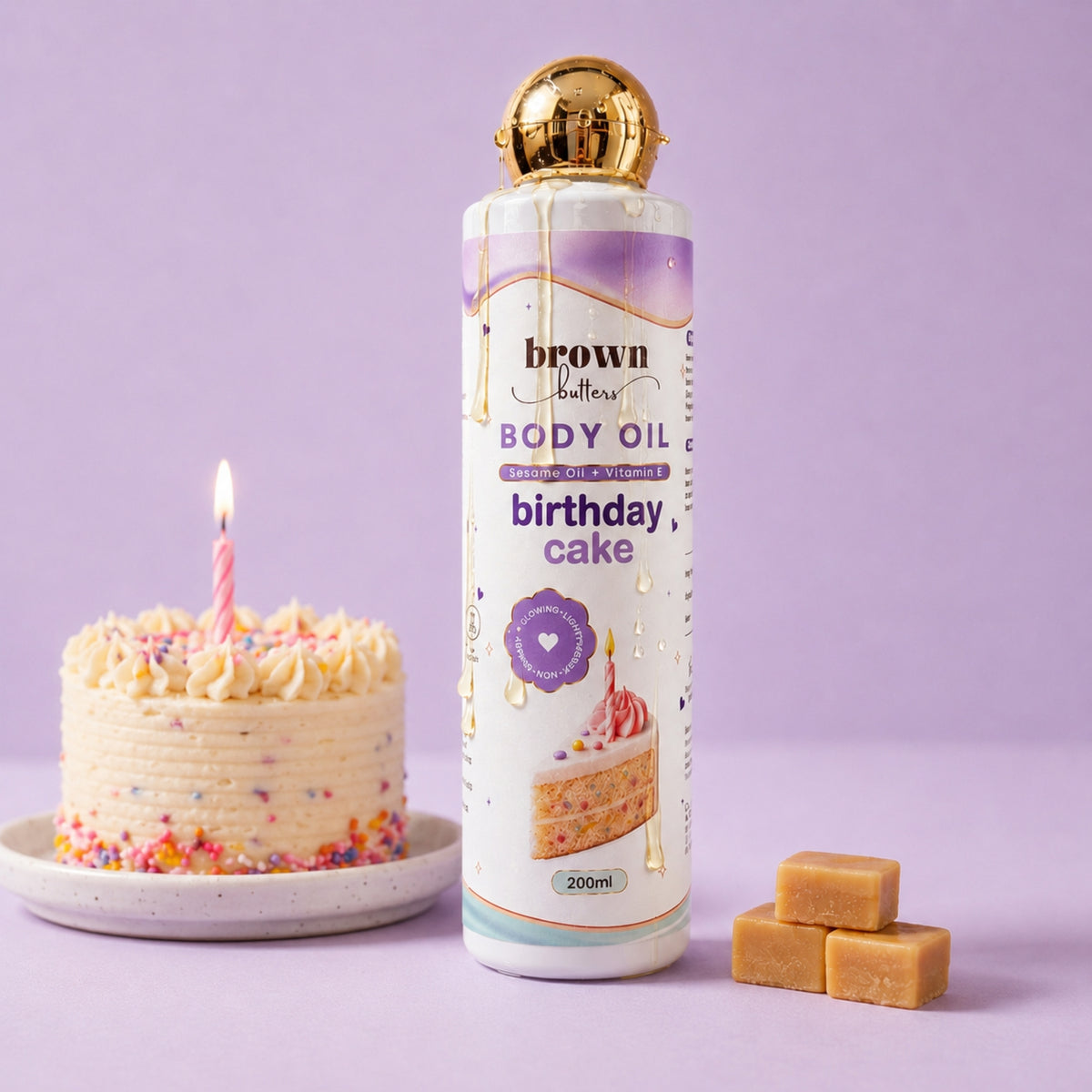 Brown Butters Birthday Cake Body Oil bottle with gold cap on lavender, styled with a small cake and caramel cubes