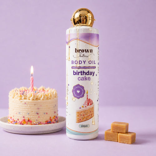Brown Butters Birthday Cake Body Oil bottle with gold cap on lavender, styled with a small cake and caramel cubes