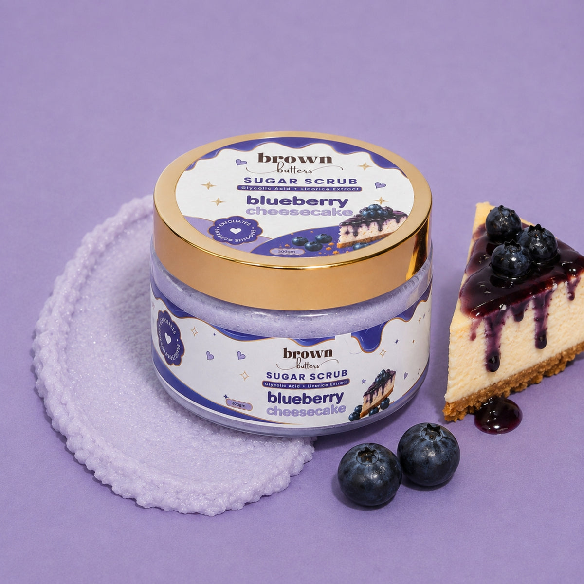 Brown Butters Blueberry Cheesecake Body Scrub jar with gold lid on lavender cloth with cheesecake slice and blueberries