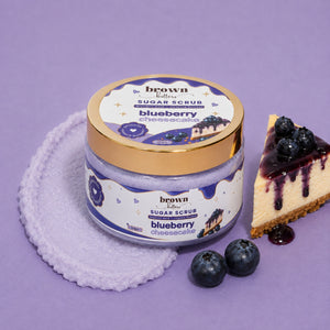 Brown Butters Blueberry Cheesecake Body Scrub jar with gold lid on lavender cloth with cheesecake slice and blueberries