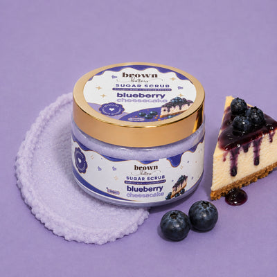 Brown Butters Blueberry Cheesecake Body Scrub jar with gold lid on lavender cloth with cheesecake slice and blueberries