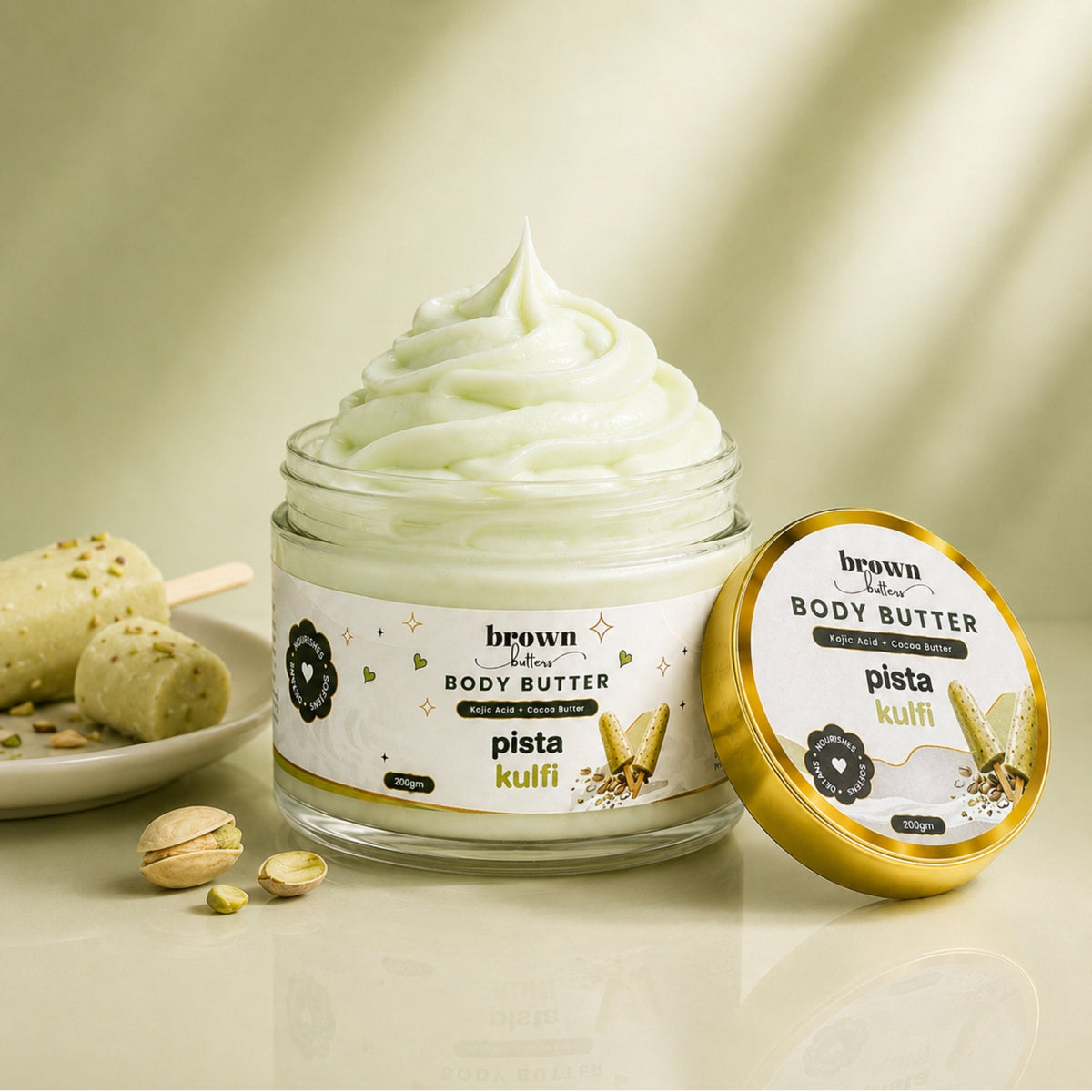 Brown Butters Pista Kulfi Body Butter jar of whipped pale green cream with its lid and pistachios on light beige