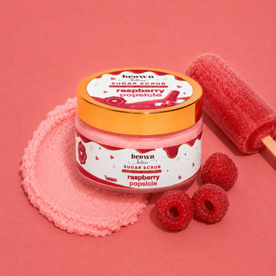 Brown Butters Raspberry Popsicle Body Scrub jar on pink with a scrub smear, fresh raspberries and a red popsicle