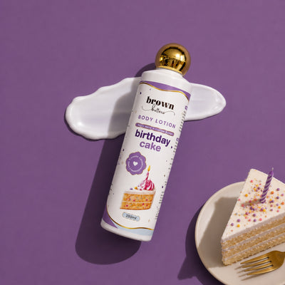 Brown Butters Birthday Cake Body Lotion bottle with gold cap on purple with a lavender smear and a cake slice