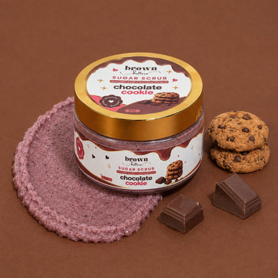 Brown Butters Chocolate Cookie Body Scrub jar on a brown scrub texture with chocolate chip cookies and chocolate
