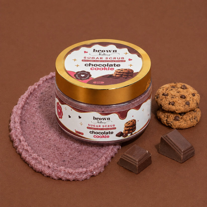 Brown Butters Chocolate Cookie Body Scrub jar on a brown scrub texture with chocolate chip cookies and chocolate