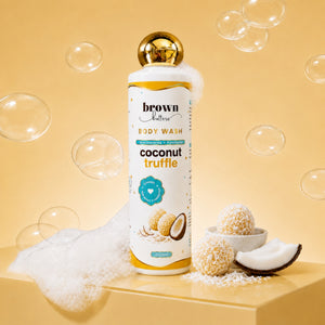 Brown Butters Coconut Truffle Body Wash bottle on a yellow pedestal with bubbles, foam and coconut props