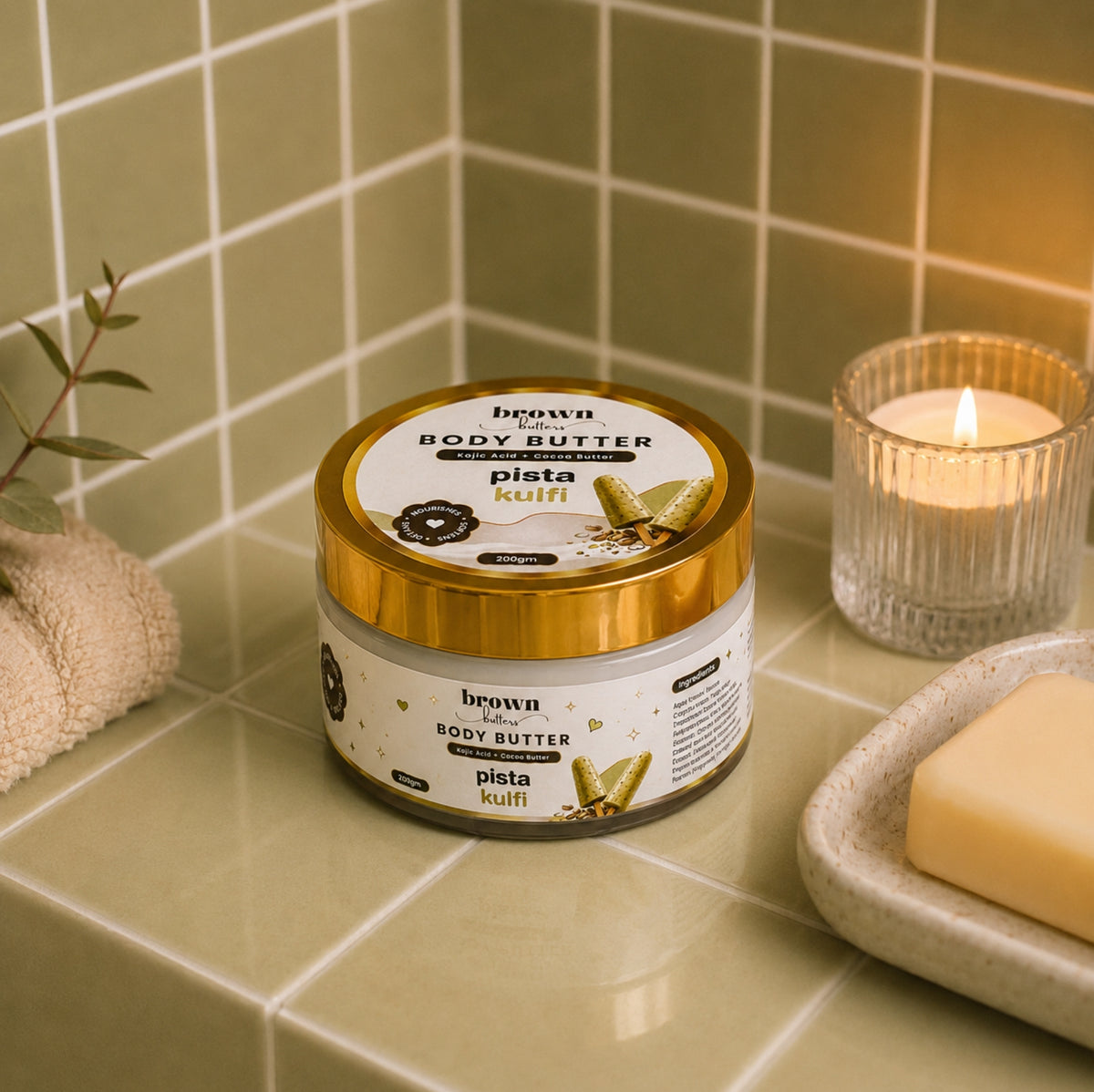 Brown Butters Pista Kulfi Body Butter jar on a green tiled bathroom counter with a lit candle, towel and soap