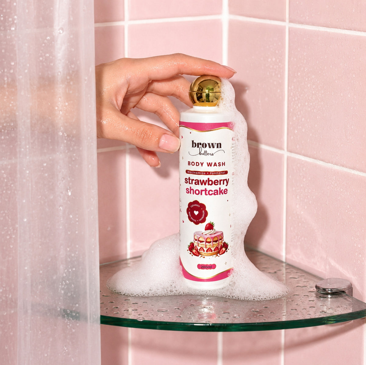 Brown Butters Strawberry Shortcake Body Wash bottle held in foam on a glass shelf against a pink tiled wall