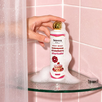 Brown Butters Strawberry Shortcake Body Wash bottle held in foam on a glass shelf against a pink tiled wall