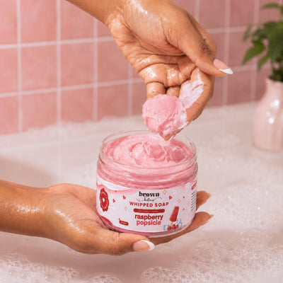 Brown Butters Raspberry Popsicle Whipped Soap scooped onto a hand in a pink tiled bathroom with bubbles