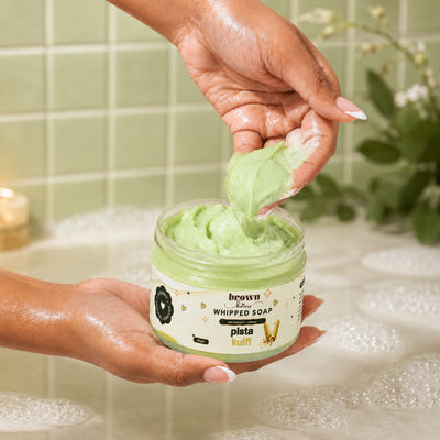 Brown Butters Pista Kulfi Whipped Soap scooped from an open jar in a tiled bathroom with bubbles and a candle