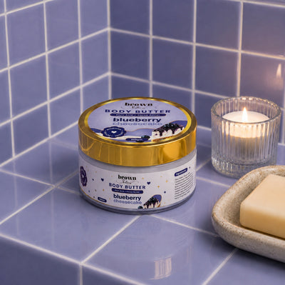 Brown Butters Blueberry Cheesecake Body Butter jar on a blue tiled surface with a lit candle and a bar of soap