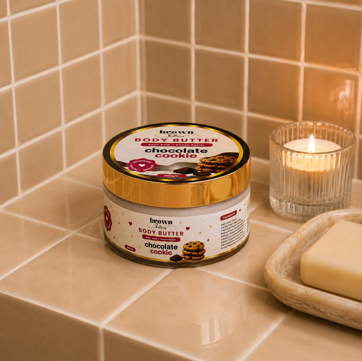 Brown Butters Chocolate Cookie Body Butter jar on a beige tiled bathroom counter with a lit candle and soap