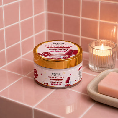 Brown Butters Raspberry Popsicle Body Butter jar on a pink tiled bathroom counter with a lit candle and soap
