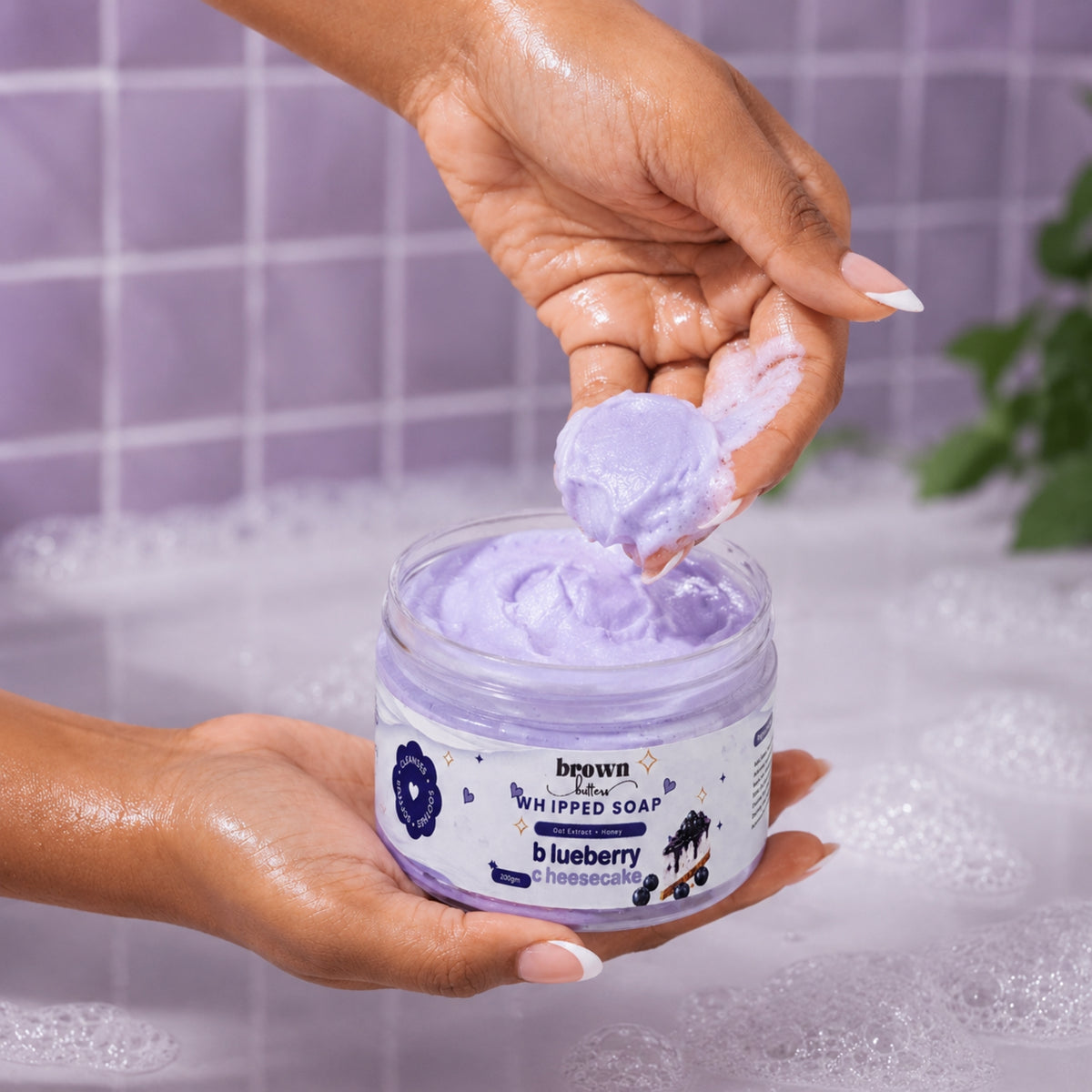 Brown Butters Blueberry Cheesecake Whipped Soap scooped by hand against a purple tiled wall and bubbly bath