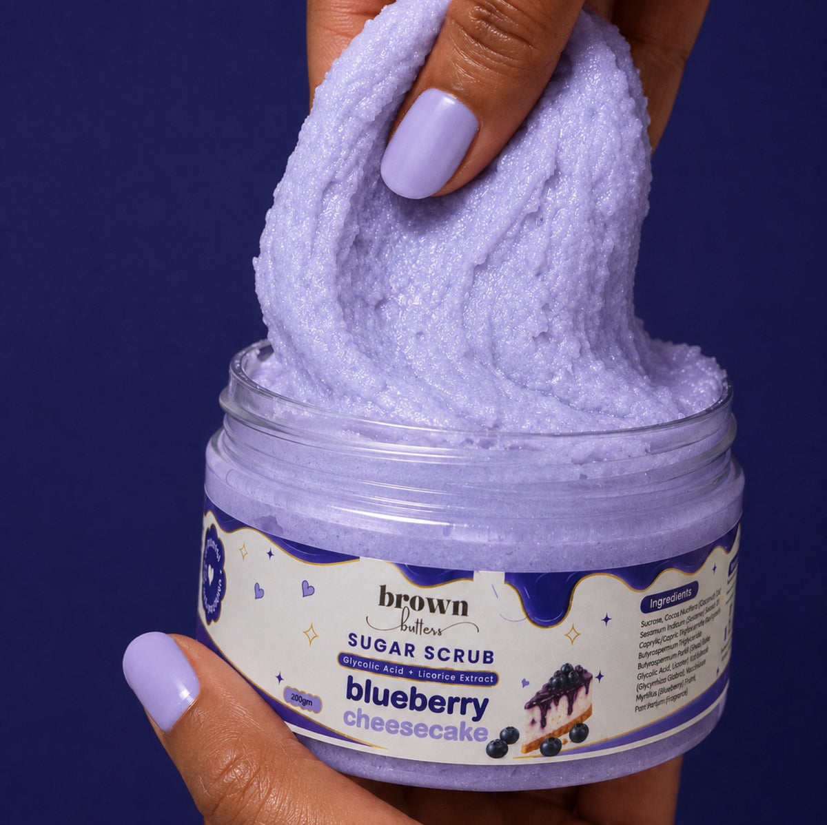 Brown Butters Blueberry Cheesecake Body Scrub stretched from its clear jar by a hand on a deep purple background
