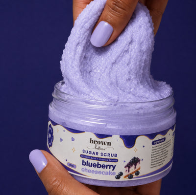 Brown Butters Blueberry Cheesecake Body Scrub stretched from its clear jar by a hand on a deep purple background