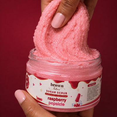 Brown Butters Raspberry Popsicle Body Scrub stretched from its clear jar by a hand on a dark red background