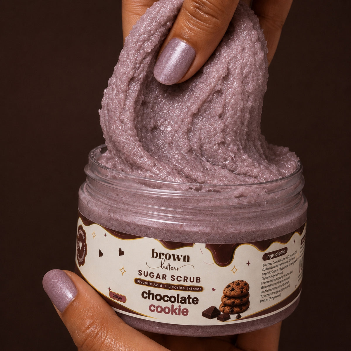 Brown Butters Chocolate Cookie Body Scrub shimmery scrub stretched from its clear jar by a hand on dark brown