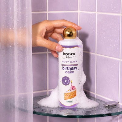 Brown Butters Birthday Cake Body Wash bottle with gold cap held on a foamy glass shelf, lavender tiles behind