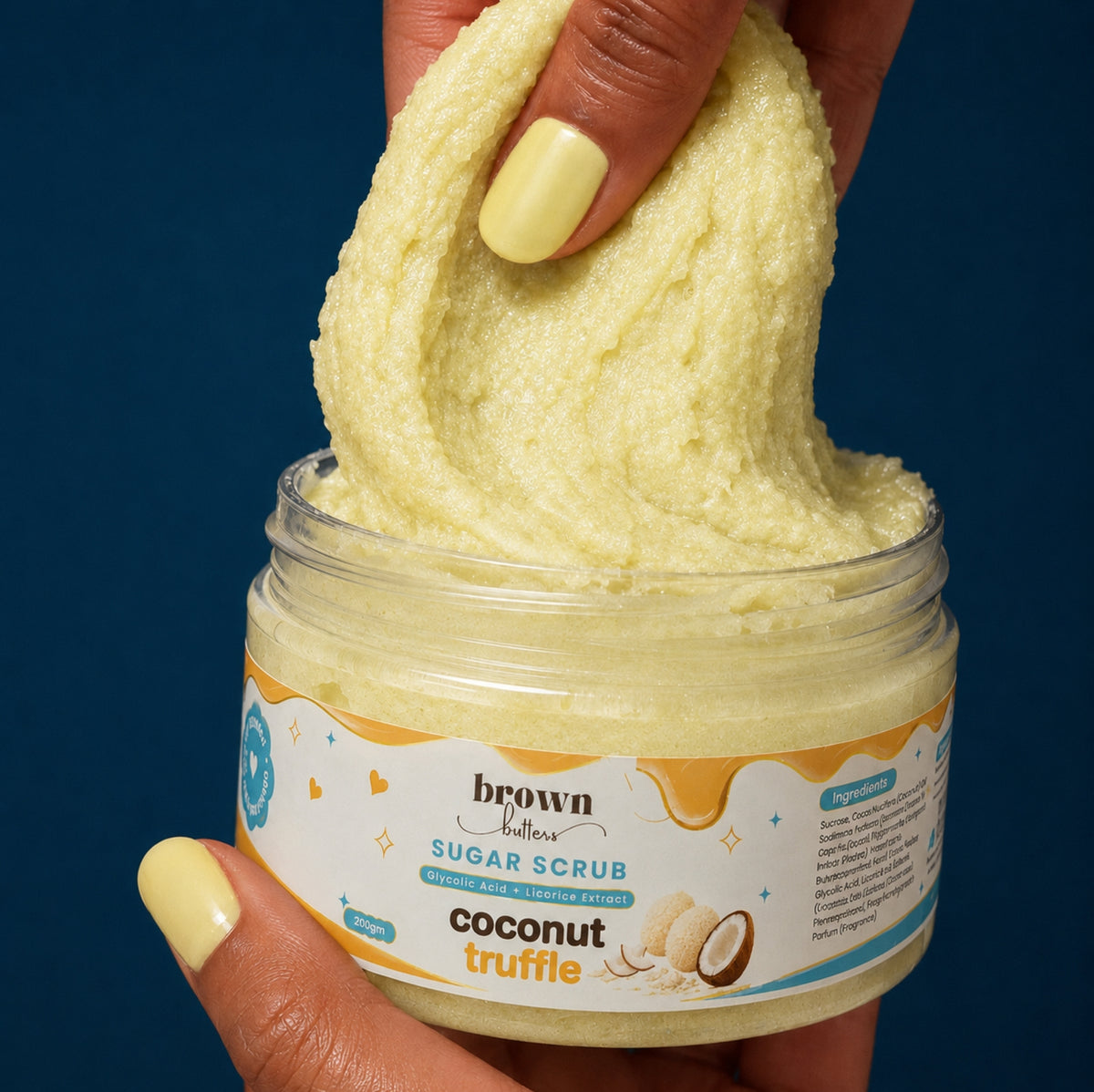 Brown Butters Coconut Truffle Body Scrub lifted in a creamy pale yellow swirl from its jar by a hand on dark blue