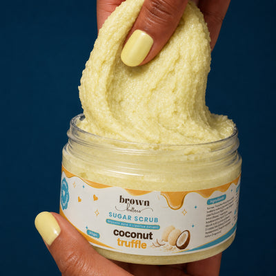 Brown Butters Coconut Truffle Body Scrub lifted in a creamy pale yellow swirl from its jar by a hand on dark blue
