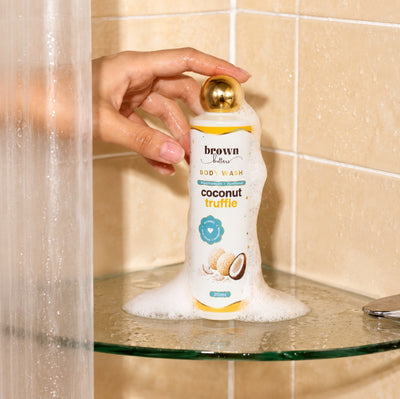 Brown Butters Coconut Truffle Body Wash bottle held with white foam dripping onto a glass shelf in a tiled bath