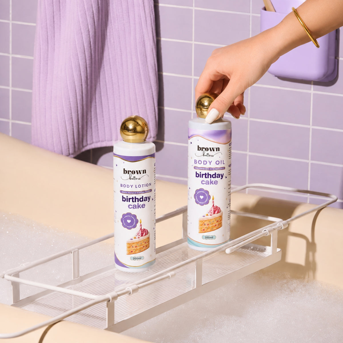 Brown Butters Birthday Cake lotion and oil on a bath rack in a bubbly tub, a hand reaching for the oil.