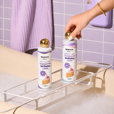 Brown Butters Birthday Cake lotion and oil on a bath rack in a bubbly tub, a hand reaching for the oil.