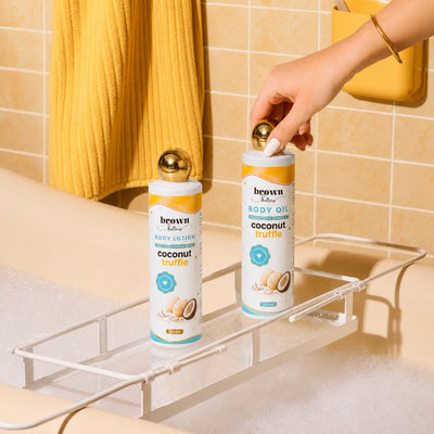 Brown Butters Coconut Truffle lotion and oil on a white bath rack in a tub, a hand reaching for a bottle.